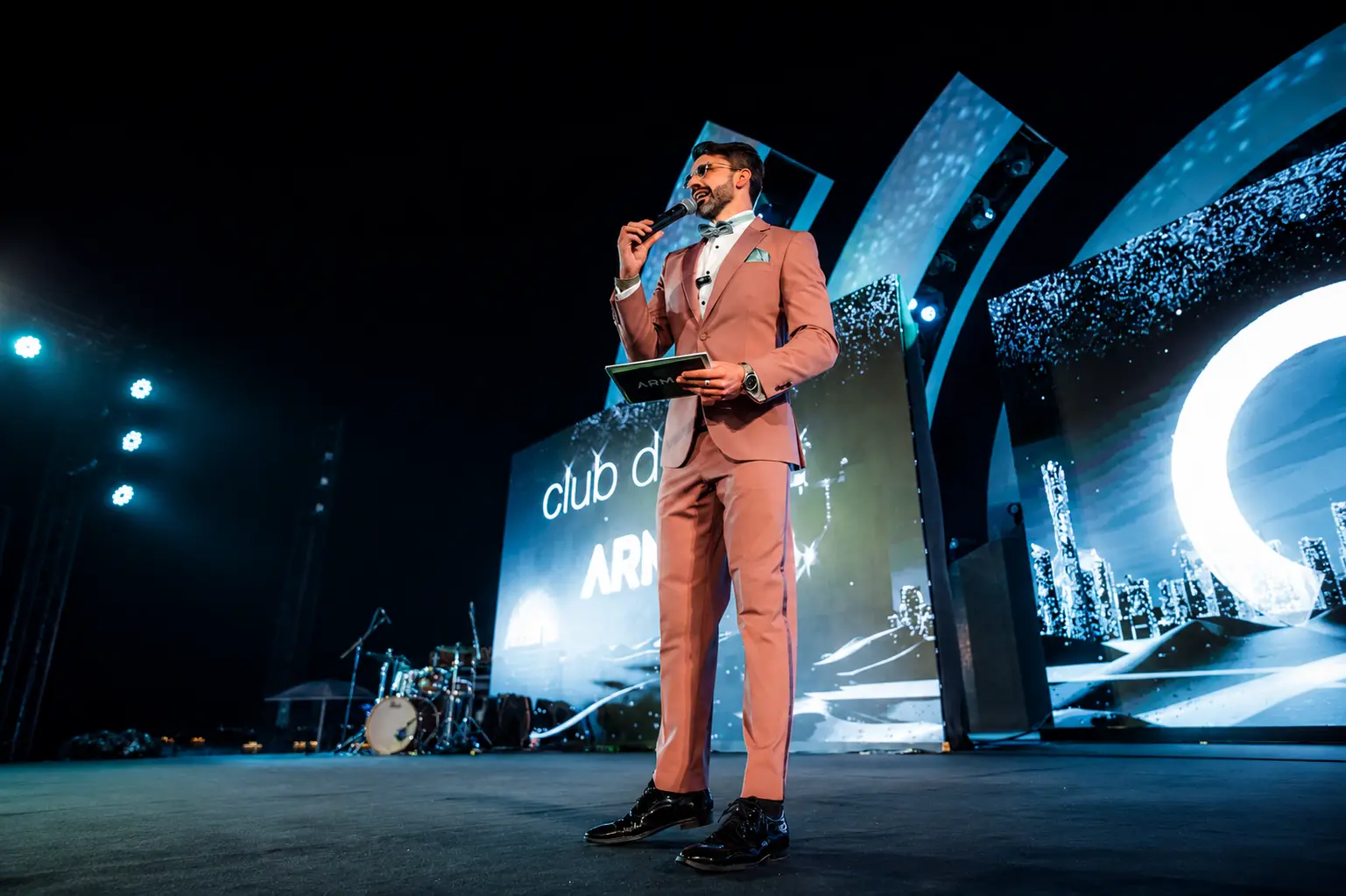 Professional MC hosting a luxury corporate awards event in Dubai, speaking on stage with a microphone in front of large LED screens.
