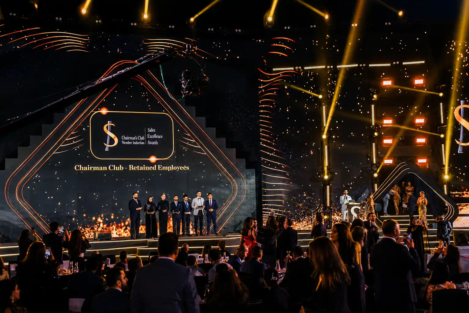 Luxury corporate awards ceremony in Dubai featuring a large illuminated stage, professional event MC, award recipients, dramatic lighting effects, and an audience celebrating during a prestigious gala event.