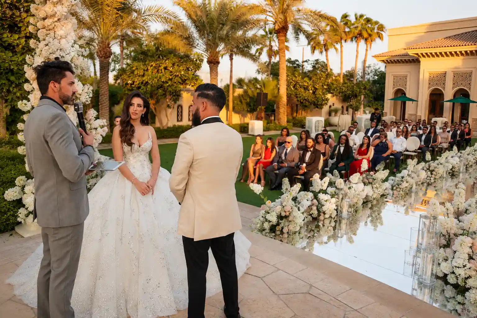 Outdoor luxury wedding ceremony in Dubai with a bride and groom standing before their celebrant, surrounded by guests, palm trees and elegant white floral decor.