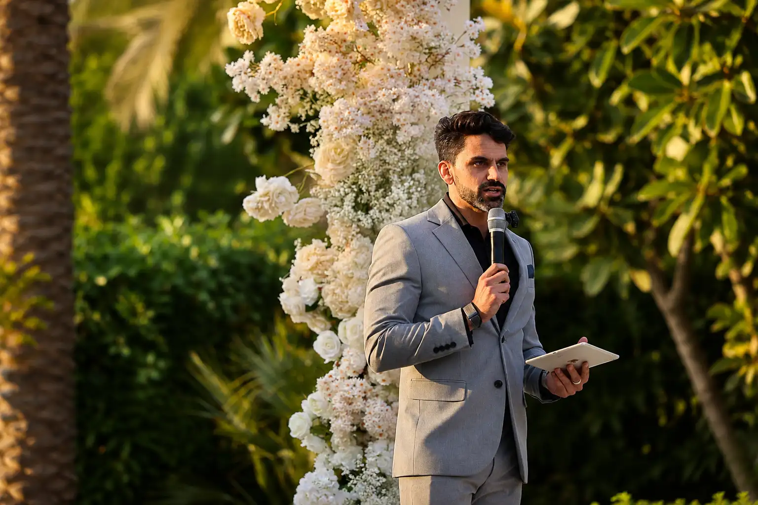 Dubai wedding MC and celebrant speaking into a microphone during an elegant outdoor garden ceremony with white floral decor.