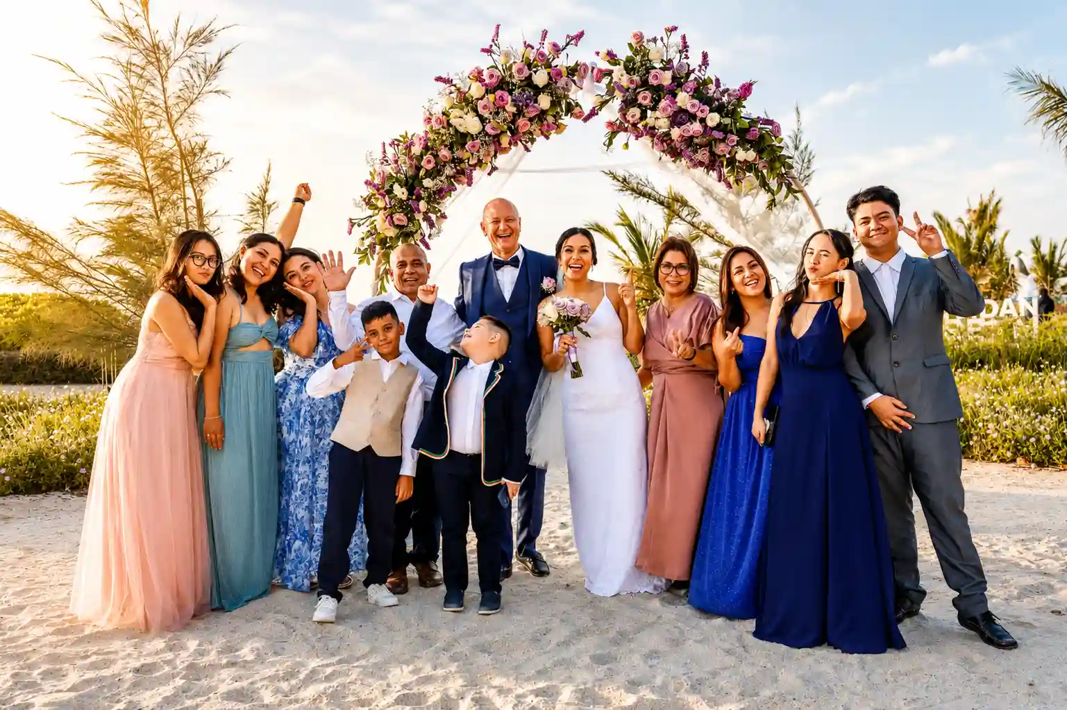 Happy bride and groom posing with family and friends beneath a floral wedding arch on a sandy beach during a joyful outdoor wedding celebration.
